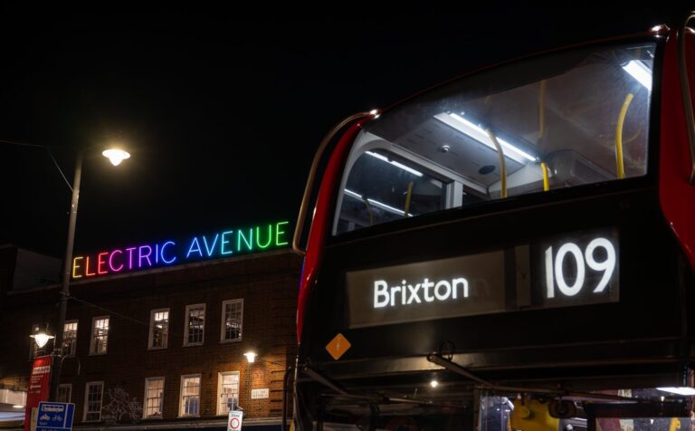 The Electric Avenue neon sign glowing in rainbow colours at night, with a red double-decker bus routed to Brixton passing in front