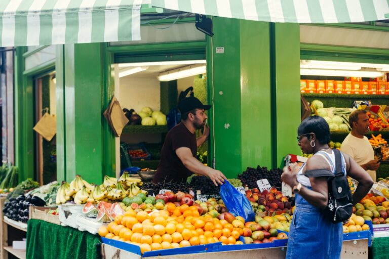 A Brixton market greengrocer's stall piled with oranges, bananas, mangoes and grapes under a striped green-and-white canopy