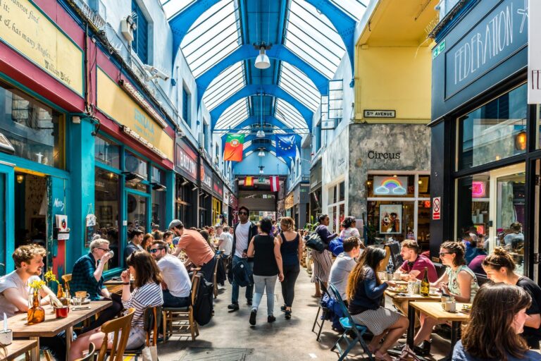Diners at outdoor cafe tables along the covered arcade inside Brixton Village, with the arched glass roof above