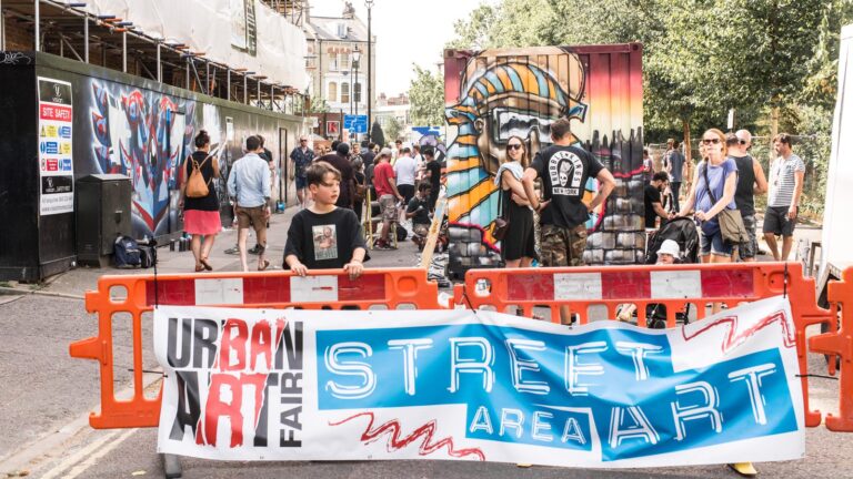 Crowds behind a street-art festival banner at Brixton's Urban Art Fair, with a large spray-painted mural in the background
