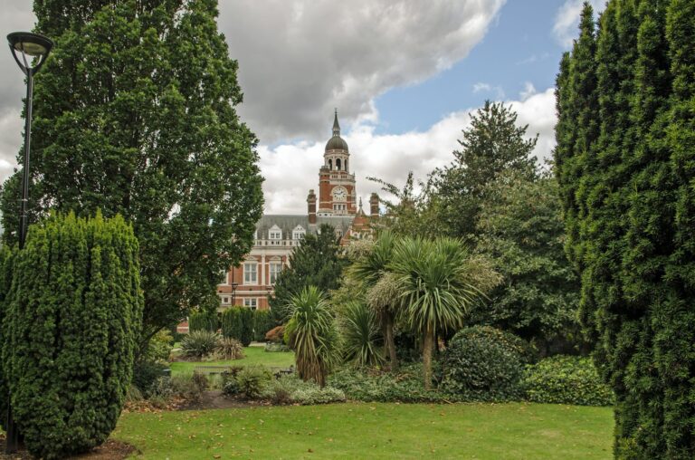 The red-brick clocktower of Croydon Town Hall framed by cypress trees and flowering shrubs in Queens Gardens on a cloudy afternoon