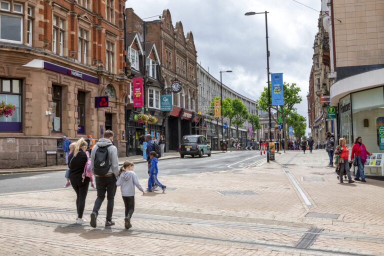 Pedestrians walking across the paved tramway on Croydon High Street, with Victorian shop fronts and hanging banners along the traffic-free stretch