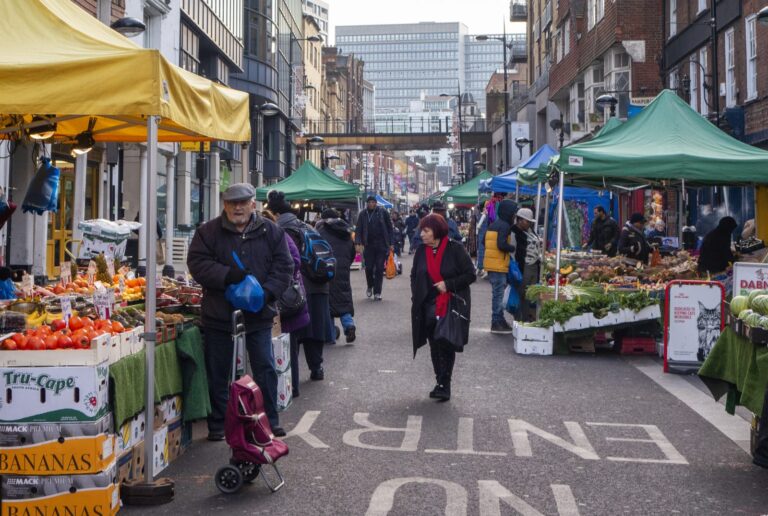 Shoppers walking between fruit and vegetable stalls on Surrey Street Market, with boxes of bananas in the foreground and a modern office block rising at the far end of the street