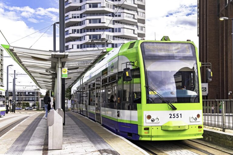 A green Tramlink tram, number 2551, pulling into a modern tram stop in central Croydon, with a curved-balcony residential tower rising behind
