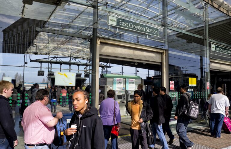 Commuters walking in and out of East Croydon Station under its modern glass canopy, with the blue 'East Croydon Station' signage overhead