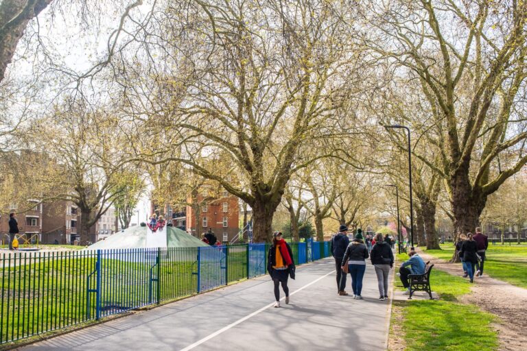 Walkers, cyclists and dog owners along the tarmac path beneath the plane trees on London Fields, with a children's playground fenced in blue on the left