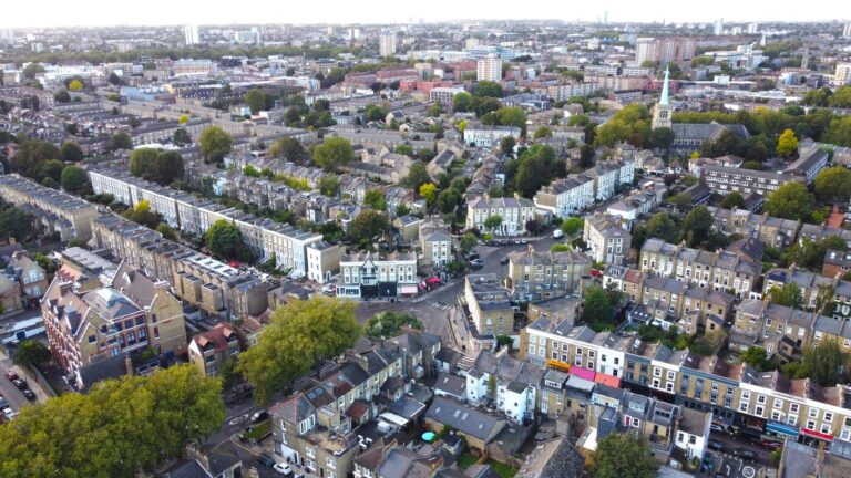 Aerial view over Hackney's Victorian terraces and high-street pubs, with church spires and distant City and Docklands towers on the horizon