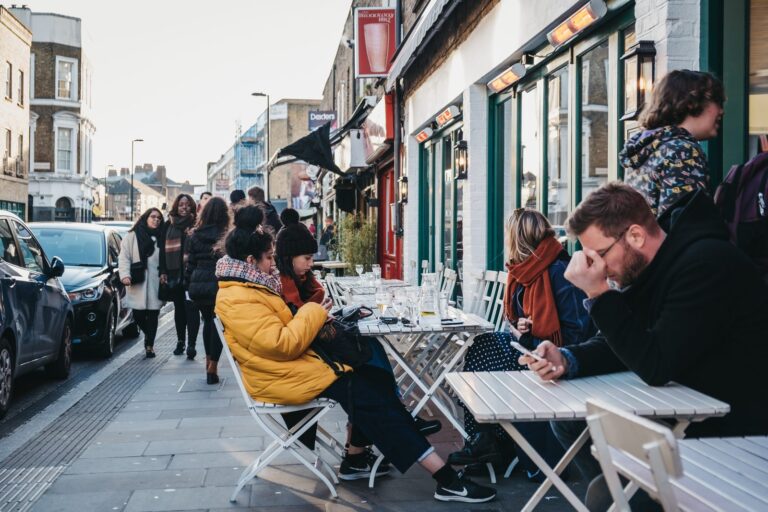 Diners at outdoor pavement cafe tables on Broadway Market on a winter weekend, with heat lamps overhead and pedestrians passing the shopfronts