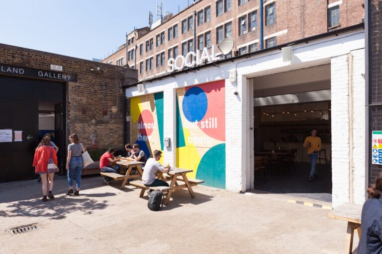 Diners at picnic benches in Copeland Park's courtyard off Rye Lane, with the Land Gallery sign and a colourful painted mural behind them