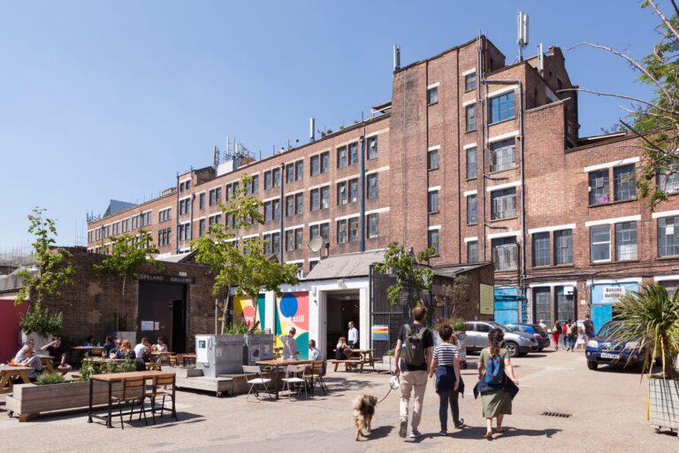 The Bussey Building's red-brick Victorian facade rises above the outdoor tables, mural wall and dog walkers passing through the Copeland Park yard
