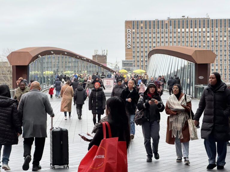 Commuters and shoppers crossing the Stratford Station footbridge above the platforms on a weekday, winter coats and Westfield shopping bags visible
