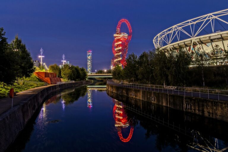 The red steel lattice of the ArcelorMittal Orbit and the lit rim of the London Stadium reflected in the still water of the Lee Navigation at night