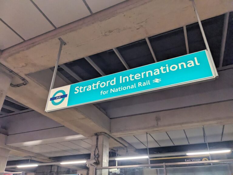 A DLR platform sign at Stratford International reading 'Stratford International for National Rail', with the East Village exit signage visible below