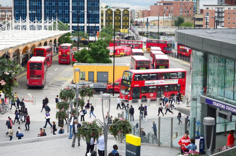 Red double-decker London buses at the Stratford bus interchange, with the glass Stratford Station entrance on the right and pedestrians crossing the plaza