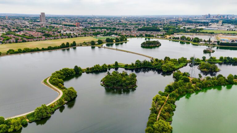 Aerial view of Walthamstow Wetlands showing the chain of reservoirs and tree-covered islands, with the east London skyline and distant towers on the horizon