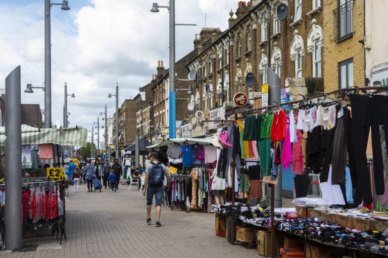 Clothing stalls and pavement shoppers along Walthamstow High Street market, with Victorian shop parades on the right and a 'Two Pound' sign on a stall to the left