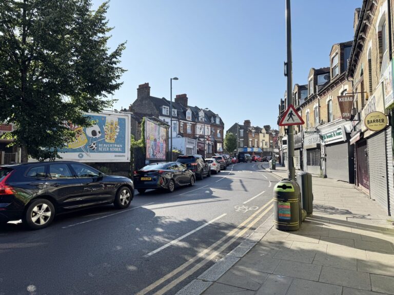 Cars lined up along Walthamstow's Hoe Street on a sunny morning, with a barber, kebab shop and a painted street mural on the left under a clear blue sky