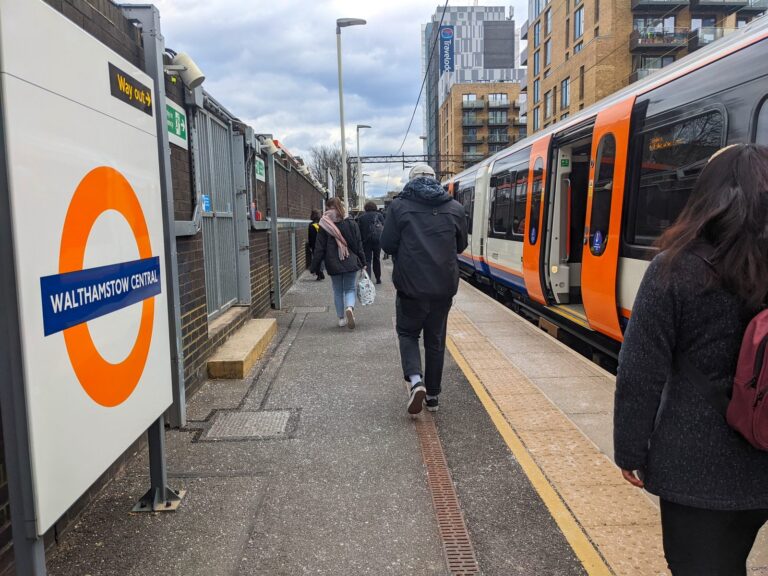 An orange-liveried London Overground train at Walthamstow Central platform, with commuters walking past a blue 'Walthamstow Central' Underground roundel sign